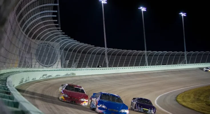Three race cars, one red, one blue, and one black, compete closely on a lit oval track at night.