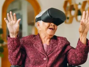 An elderly woman wearing a virtual reality headset, smiling and gesturing with her hands, seated in a cozy living room setting.