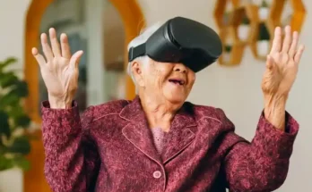 An elderly woman wearing a virtual reality headset, smiling and gesturing with her hands, seated in a cozy living room setting.