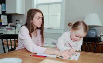A woman and child sit at a wooden table engaged in an art project, surrounded by colorful pencils and paper in a bright room.