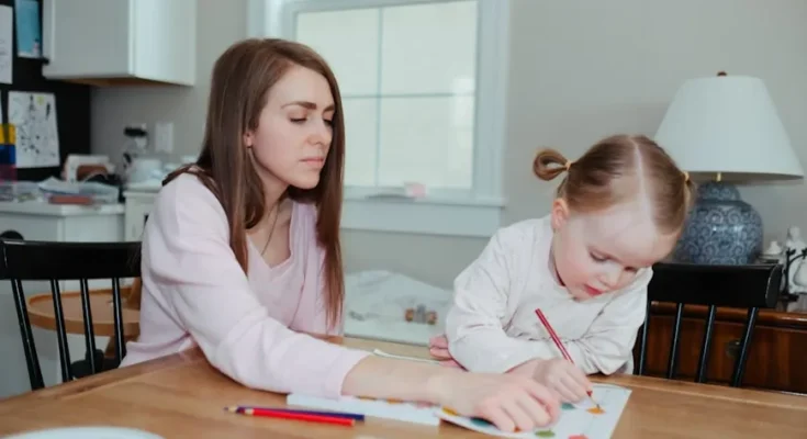 A woman and child sit at a wooden table engaged in an art project, surrounded by colorful pencils and paper in a bright room.