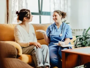 A healthcare professional in scrubs discusses care with a patient sitting on a couch in a cozy, well-lit living room setting.