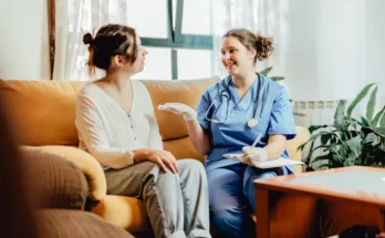 A healthcare professional in scrubs discusses care with a patient sitting on a couch in a cozy, well-lit living room setting.