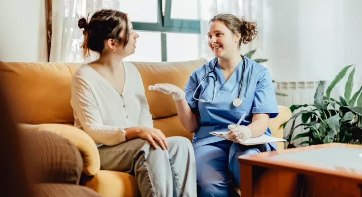 A healthcare professional in scrubs discusses care with a patient sitting on a couch in a cozy, well-lit living room setting.