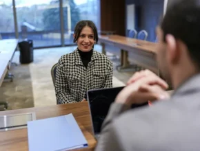 A woman in a houndstooth jacket sits across from a man in a suit at a meeting table, engaged in a conversation.