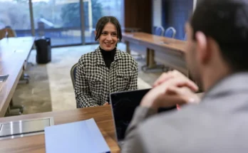 A woman in a houndstooth jacket sits across from a man in a suit at a meeting table, engaged in a conversation.