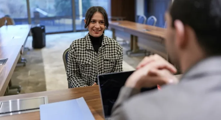 A woman in a houndstooth jacket sits across from a man in a suit at a meeting table, engaged in a conversation.