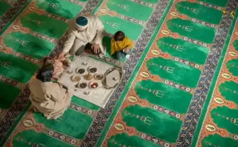 An overhead view of two adults and a child gathered around a plate of traditional food on patterned green prayer mats.