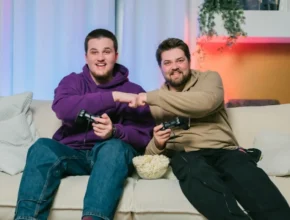 Alt Text: Two friends share a moment on a couch, gaming together and bumping fists, with a bowl of popcorn between them.