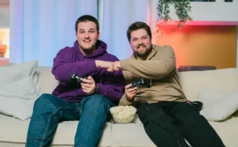 Alt Text: Two friends share a moment on a couch, gaming together and bumping fists, with a bowl of popcorn between them.