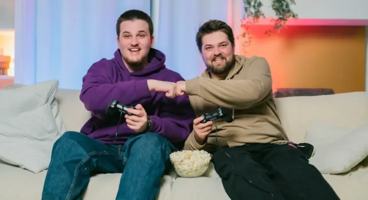 Alt Text: Two friends share a moment on a couch, gaming together and bumping fists, with a bowl of popcorn between them.