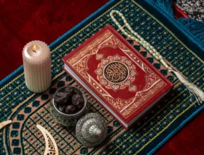 A beautifully ornate Quran rests on a colorful prayer mat alongside a lit candle, dates, and prayer beads, evoking a serene atmosphere.