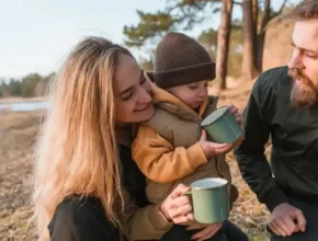 A woman and two individuals enjoy a cozy outdoor moment with a child, sipping from green mugs by a serene lakeside.