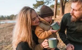 A woman and two individuals enjoy a cozy outdoor moment with a child, sipping from green mugs by a serene lakeside.
