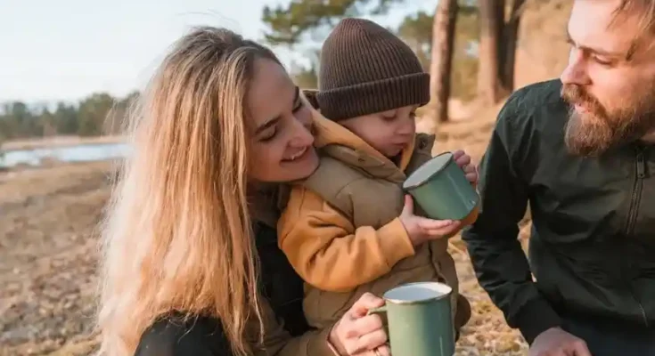 A woman and two individuals enjoy a cozy outdoor moment with a child, sipping from green mugs by a serene lakeside.