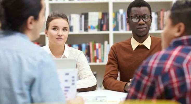 A group of four people engaged in discussion around a table, surrounded by shelves filled with colorful books in a cozy environment.