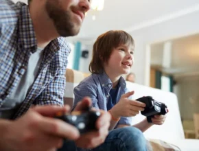 A man and a boy sit side by side on a couch, engrossed in video gaming, each holding a game controller.