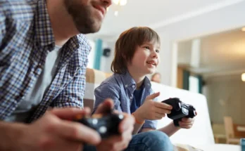 A man and a boy sit side by side on a couch, engrossed in video gaming, each holding a game controller.