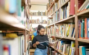 A person stands between bookshelves in a library, reading a book while surrounded by colorful, neatly organized books.