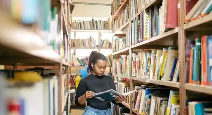 A person stands between bookshelves in a library, reading a book while surrounded by colorful, neatly organized books.