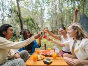 A group celebrating with drinks and food, surrounded by nature. A picnic table features watermelon and cookies.