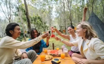 A group celebrating with drinks and food, surrounded by nature. A picnic table features watermelon and cookies.