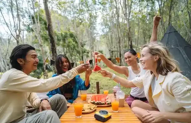 A group celebrating with drinks and food, surrounded by nature. A picnic table features watermelon and cookies.