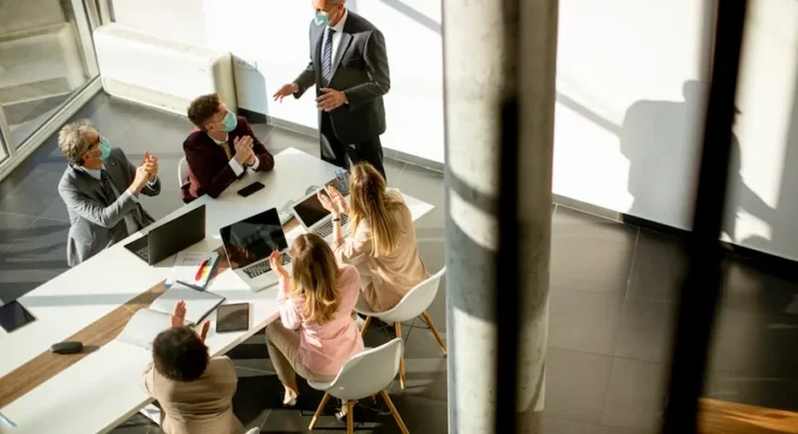 A diverse group of professionals in masks engaged in a lively meeting at a modern conference table with laptops and notepads.