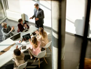 A group of professionals in a modern conference room engage in discussion, with laptops and a presentation setup on the table.