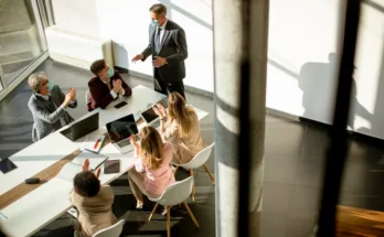 A group of professionals in a modern conference room engage in discussion, with laptops and a presentation setup on the table.