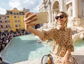A person in a polka dot dress takes a selfie at the Trevi Fountain, surrounded by a bustling crowd and classic Roman architecture.
