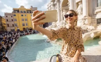 A person in a polka dot dress takes a selfie at the Trevi Fountain, surrounded by a bustling crowd and classic Roman architecture.