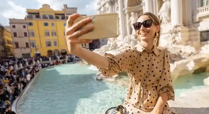 A person in a polka dot dress takes a selfie at the Trevi Fountain, surrounded by a bustling crowd and classic Roman architecture.