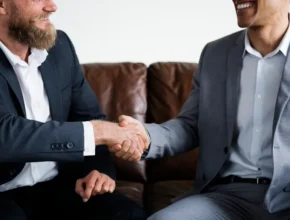 Two businessmen in suits shake hands over a brown leather couch, conveying a sense of agreement and collaboration.