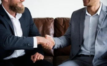 Two businessmen in suits shake hands over a brown leather couch, conveying a sense of agreement and collaboration.