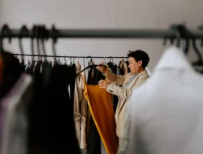 A person in a stylish jacket examines a vibrant yellow dress on a clothing rack filled with various garments in a bright room.