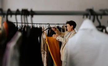 A person in a stylish jacket examines a vibrant yellow dress on a clothing rack filled with various garments in a bright room.