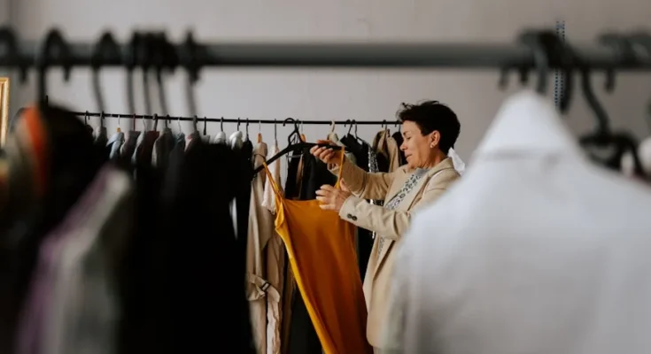 A person in a stylish jacket examines a vibrant yellow dress on a clothing rack filled with various garments in a bright room.