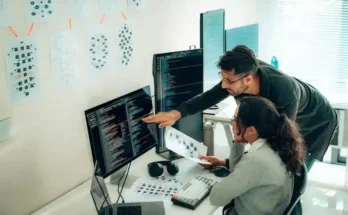 A woman and a man collaborate at a desk surrounded by monitors displaying code and project images, with notes pinned on the wall.