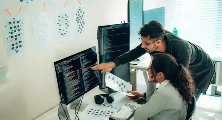 A woman and a man collaborate at a desk surrounded by monitors displaying code and project images, with notes pinned on the wall.