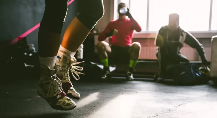 A person jumps rope in a gym, focused on their exercise; two seated individuals watch in the background, engaging in conversation.