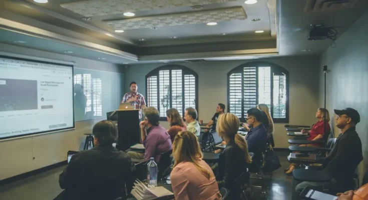 A speaker presents in a well-lit conference room, with an engaged audience attentively facing a large projection screen.