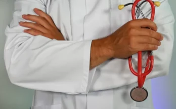 A doctor in a white lab coat crosses arms, holding a red stethoscope, symbolizing professionalism and healthcare expertise.