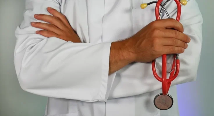 A doctor in a white lab coat crosses arms, holding a red stethoscope, symbolizing professionalism and healthcare expertise.