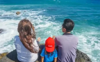 A family of three sits on a rocky ledge by the sea, gazing at the waves and enjoying the sunny, vibrant ocean view.