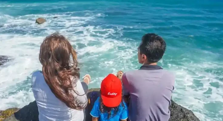 A family of three sits on a rocky ledge by the sea, gazing at the waves and enjoying the sunny, vibrant ocean view.