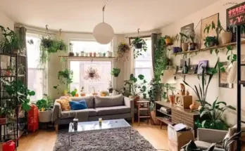 A cozy living room filled with lush plants, featuring a gray sofa, a glass coffee table, and bright natural light from large windows.