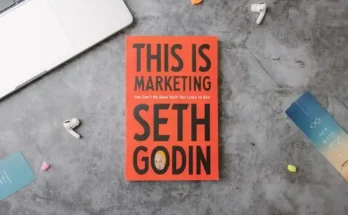 A laptop, earbuds, and colorful sticky notes surround the book "This Is Marketing" by Seth Godin, titled in bold black on a red cover.