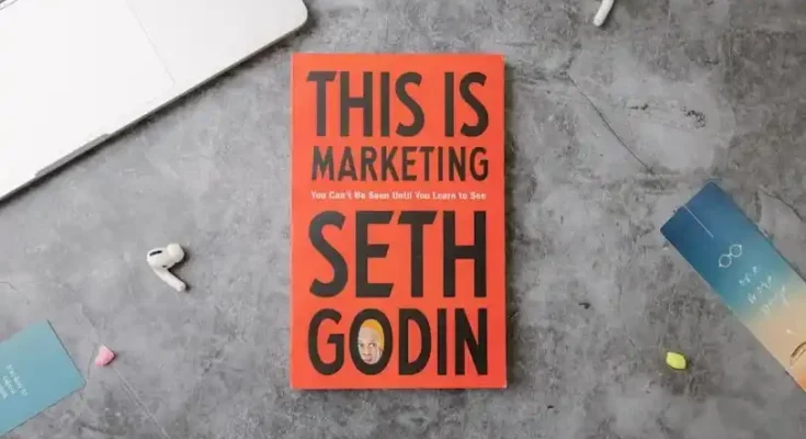 A laptop, earbuds, and colorful sticky notes surround the book "This Is Marketing" by Seth Godin, titled in bold black on a red cover.