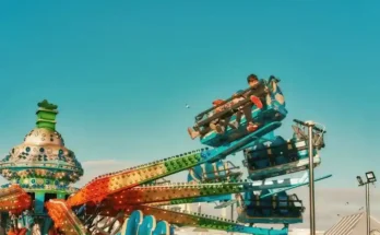 Two children enjoy a thrill ride at a carnival, suspended high against a bright blue sky filled with a few scattered clouds.